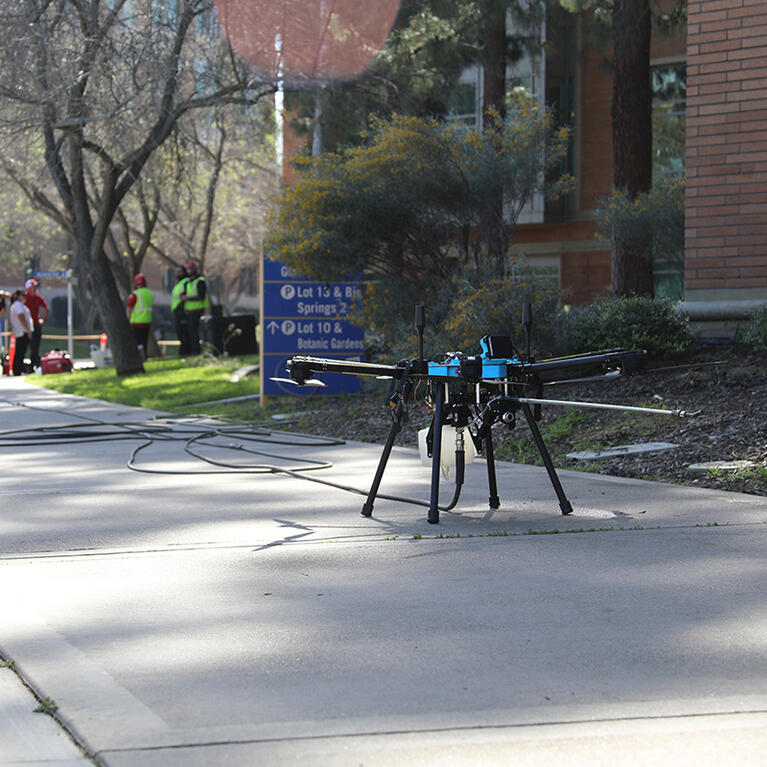 A black drone sits on the sidewalk next to a brick building with a van in the background